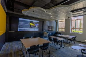 An image showing the Social Media Lab with a website showing large on the wall of monitors. The room has severall large tables with chairs around them.