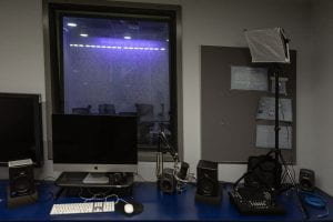 An image of the podcast studio from the inside of the edit bay. Also showing the edit bay's iMac, TV, microphone and speakers.