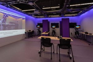 An image of the IML, purple LEDs shine down from the ceiling. There are three VR headsets next to three computers on carts separated by partitions. The room's projector is displayed a website on the wall.